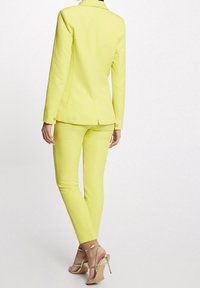 Vrouw in een helder gele op maat gemaakte pak met lange mouwen en bijpassende smalle broek, gecombineerd met metallic gouden hoge hakken sandalen.