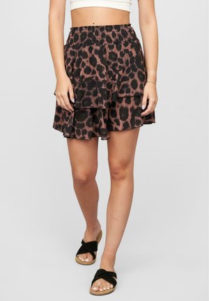 Vrouw in een bruin en zwart luipaardprint gelaagde mini-rok, witte crop top en zwarte sandalen die op een grijze achtergrond staat.