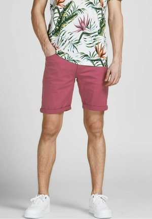 Różowe bawełniane szorty z podwiniętymi nogawkami, noszone z białą koszulą w tropikalny wzór. Model stoi swobodnie, ręce w kieszeniach, ubrany w białe trampki.