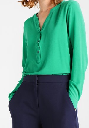 Femme portant un chemisier vert à manches longues avec des boutons et un pantalon taille haute bleu marine, mains dans les poches.