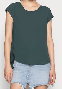 Femme portant un haut à manches courtes vert foncé et une jupe en denim bleu clair avec un ourlet effiloché et une fente à l'avant.