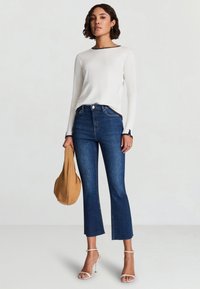 Top bianco a maniche lunghe con rifiniture blu navy, jeans blu scuro aderenti con orlo corto, sandali con tacco trasparente e una borsa color tan.