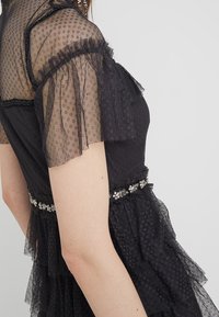 Mujer vistiendo un vestido negro transparente con lunares en capas, con mangas abullonadas y un cinturón de cintura adornado con rhinestones, vista de lado.