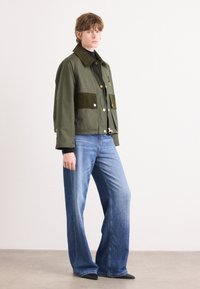 Olijfgroene jas met een stevige textuur, voorzien van twee fluwelen voorzakken, gouden hardware en een kraag, gecombineerd met blauwe uitlopende jeans.