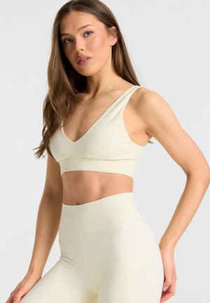 Jonge vrouw die een bijpassende lichtbeige sportbeha en hooggetailleerde leggings draagt, staat met één hand op de heup tegen een witte achtergrond.