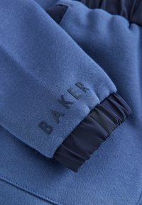Manche en tissu bleu avec poignet élastique noir et lettres noires en relief formant le mot "BAKER" près du poignet.