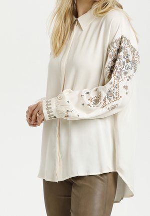 Femme portant un chemisier ample crème avec une broderie brune et grise complexe sur les manches, associée à un pantalon en cuir marron, les mains jointes.