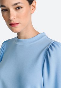 Hele sinine pehme kangast ülaosa, millel on kõrge ümar kaelus ja õlgadel voltidega pintsakvarrukad.