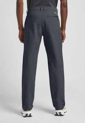 Pantaloni da uomo grigio scuro realizzati in un tessuto leggero e liscio, dotati di una tasca posteriore con zip e una vita regolabile. Accentuati da sneakers.