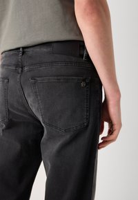 Vaqueros de mezclilla negros con un corte ajustado, que cuentan con dos bolsillos traseros, un parche de logo de cuero y un brillo sutil. Detalles de costura a lo largo de las costuras.