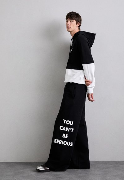 Čierna a biela mikina s kapucňou a skráteným strihom, spárovaná s voľnými čiernymi nohavicami s bielym textom: "NEMÔŽEŠ TO MISLIEŤ VÁŽNE."