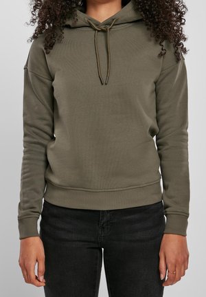 Personne portant un sweat à capuche vert olive avec cordons et un jean noir, debout avec les bras détendus le long du corps devant un fond uni.