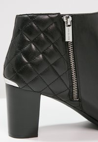 Bottine noire à talon bloc avec texture matelassée, fermeture éclair sur le côté. Fabriquée en cuir avec des accents métalliques. Fini lisse.