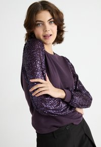 Violetinis megztinis su blizgučiais padengtomis rankovėmis, minkšta tekstūra ir apvaliu kaklu. Suderintas su juodomis kelnėmis, pabrėžiančiomis prigludusį stilių.