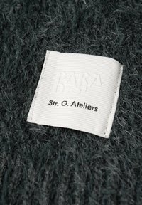 Mørkegrøn, struktureret stof med et fremtrædende hvidt label med præget tekst, der læser "PARA DEST" og "Str. O. Ateliers."