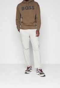 Hnědá mikina s kapucí s logem "BOSS", kombinovaná s světlejšími cargo kalhotami a vícejazyčnými teniskami s šedými, fialovými a černými akcenty.