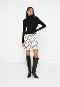 Žena stoji s rukom na kuku, nosi crnu majicu dugih rukava, bijelu suknju s cvjetnim uzorkom i crne visoke čizme s vrhom špičastim prema gore na bijeloj pozadini.