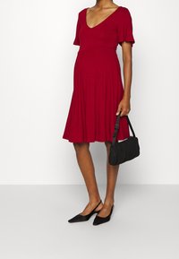 Kobieta w czerwonej sukience do kolan, z krótkimi rękawami, w czarnych butach na obcasie typu slingback, trzymająca małą czarną torebkę przy boku.