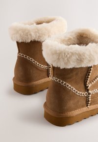 Bottes chevilles en daim marron avec un col en fausse fourrure beige, des détails cousus et des semelles en caoutchouc avec un motif de bande de roulement texturé.
