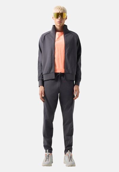 Osoba z krótkimi blond włosami nosi duże okulary przeciwsłoneczne o żółtych szkłach, szary dres, pomarańczową koszulkę i jasne buty sportowe, stoi prosto.