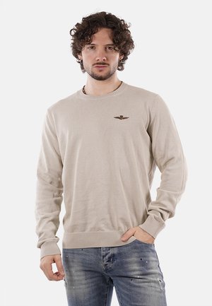 Maglione beige lavorato a maglia con collo rotondo e polsini a coste, caratterizzato da un piccolo logo ricamato sul petto, indossato con jeans di denim.