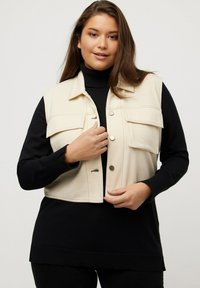 Gilet beige tagliato con quattro tasche frontali e bottoni argentati, indossato sopra un dolce a collo alto nero. Tessuto liscio con un design strutturato.