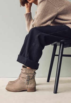 Bottes à chevilles en daim beige avec un design froissé, des accents de boucle et une texture matelassée. Portées avec un jean sombre retroussé et un pull léger.