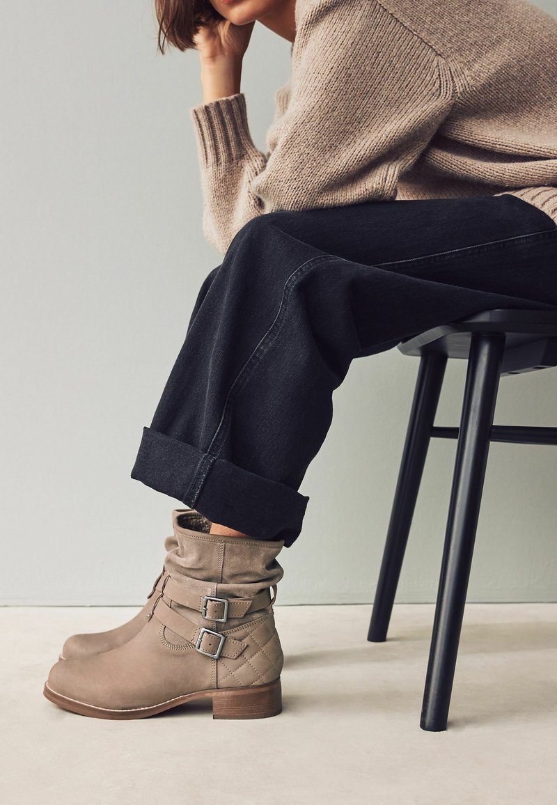 Bottes à chevilles en daim beige avec un design froissé, des accents de boucle et une texture matelassée. Portées avec un jean sombre retroussé et un pull léger.