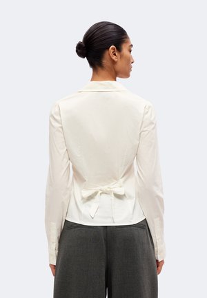 Une femme vue de dos porte un chemisier blanc noué à la taille et un pantalon gris foncé sur un fond clair et uni.