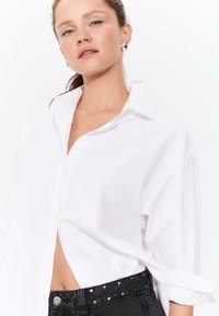 Woman wearing oversized white button-up shirt with sleeves rolled up, black studded pants, silver geometric earrings, against plain white background.