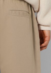 Beiget hännät, jotka sisältävät suoran lahkeeen, pehmeän kankaan ja sivutaskun. Vyötärönauha on joustava. Kermanvalkoinen neulepaita näkyy osittain.