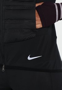 Černá sportovní bunda s designem prošívání a zipovou kapsou; obsahuje stříbrné logo Nike a strukturované textilní akcenty.