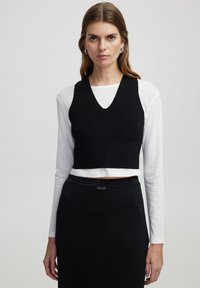 Černý řasený crop top s výstřihem do V přes bílou dlouhou košili, spárovaný s černou sukní s hladkou texturou a přiléhavým střihem.