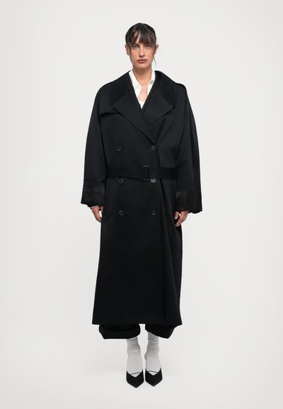 Schwarzer doppelt geöffneter Trenchcoat mit breiten Revers, tailliertem Bund und langen Ärmeln. Getragen über schwarzen Hosen und weißen Socken.