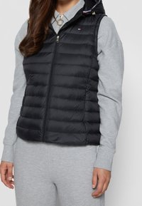 Czarna pikowana kamizelka typu puffer z zamkiem błyskawicznym z przodu, pikowanym wzorem i kapturem. Posiada złote wykończenia oraz mały detal z logo na piersi.