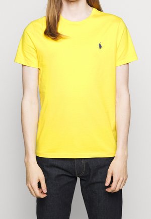 Gelbes Baumwoll-T-Shirt mit Rundhalsausschnitt, kurzen Ärmeln und einem kleinen blauen Logo auf der linken Brust. Getragen mit dunklen Jeans.