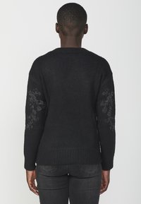 Maglione nero con maniche lunghe, orlo a coste e motivi decorativi di fiocchi di neve argentati sulle maniche. Morbido al tatto, design casual.