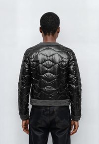 Zwarte gewatteerde jas met een glanzende afwerking, voorzien van een golvend quiltpatroon, geribde kraag en manchetten. Gestyled met een donkere spijkerbroek.
