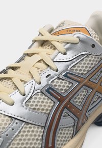 Zbliżenie beżowego sneakersa z siateczkową tkaniną, kremowymi sznurówkami, srebrnymi nakładkami oraz brązowo-niebieskim logo na boku, z napisem GEL-1130 na języku.