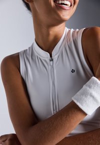 Biała bezrękawnikowa koszulka sportowa z wysokim kołnierzem i zamkiem z przodu, z małym czarnym logo. Na jednym ramieniu widoczna jest biała opaska na nadgarstek.