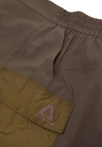 Plano detalle de unos pantalones cortos marrones con cintura elástica y un bolsillo con solapa verde que lleva el logo de Altavia en púrpura apagado.