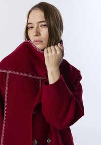 Červený vlněný kabát s vysokým límcem, který má kontrastní bílé švy, velké knoflíky a volný střih. Texturovaná tkanina s hladkým povrchem.