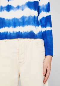 Sinivalkoinen värjäyksellä tehty neulepaita, jossa on ribbineulotut hihansuut ja helma, yhdistetty norsunluunvärisiin farkkuihin. Käsi lepää housujen päällä.