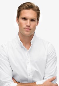 Witte lange mouwen overhemd met knoopsluiting, van strakke stof, met kraag, met een subtiele logo op de borst, gedragen met de armen gekruist.