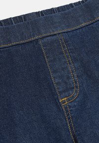 Mørkeblåt denimstof med gul kontrastsyning og detaljer med elastisk taljebånd øverst, der viser omridset af en jeanslomme og en søm.