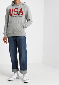 Sudadera gris claro con gráfico rojo "USA", que cuenta con un bolsillo canguro y cordones. Lleva unas jeans de mezclilla azul oscuro con los puños enrollados y zapatillas blancas.