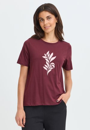 Femme portant un t-shirt bordeaux à manches courtes avec un motif abstrait de feuille rose clair, associé à un pantalon noir, debout devant un fond uni.