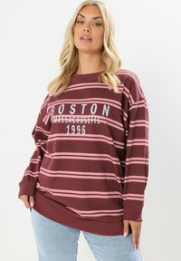 Blondynka w bordo i różowym pasiastym dresie z napisem "Boston Massachusetts 1996", w połączeniu z jasnoniebieskimi dżinsami.