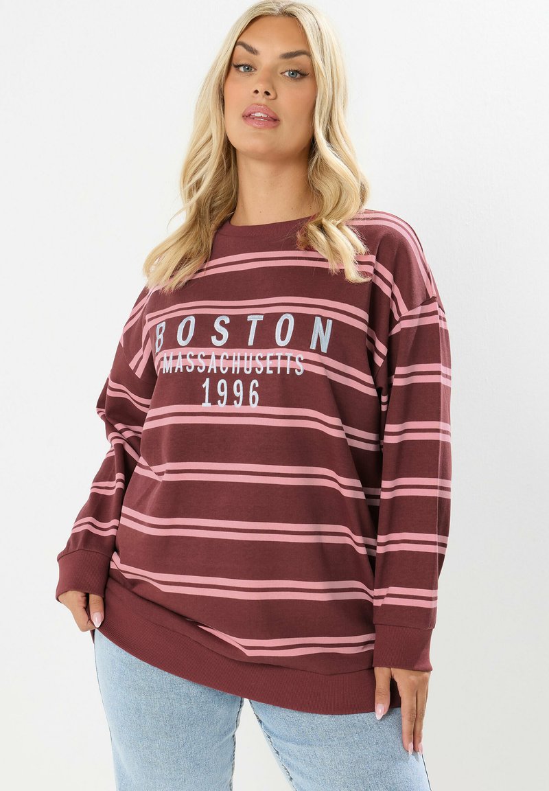 Blondynka w bordo i różowym pasiastym dresie z napisem "Boston Massachusetts 1996", w połączeniu z jasnoniebieskimi dżinsami.