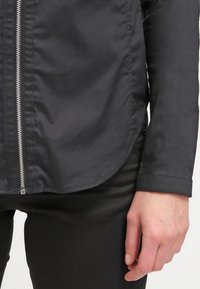 Svart lättviktsjacka med en slät textur, med en silverfärgad dragkedja framtill, rundad nederkant och knappdetalj vid ärmsluten.